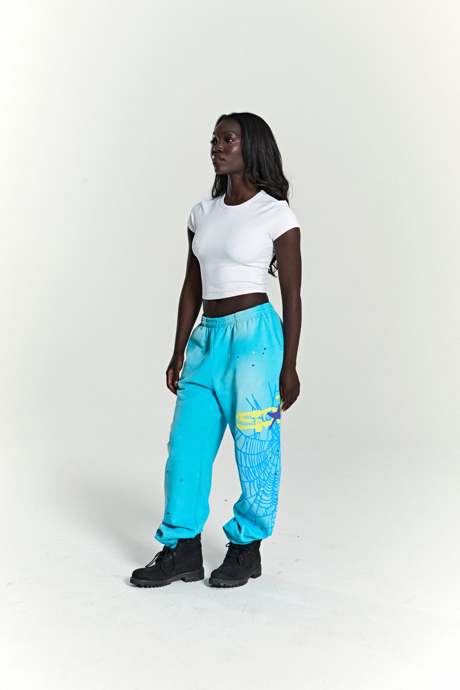 Woman wearing turquoise HEAVY WEB SWEATPANTS with SP5DER web graphic, scattered stars, and elasticized cuffs.