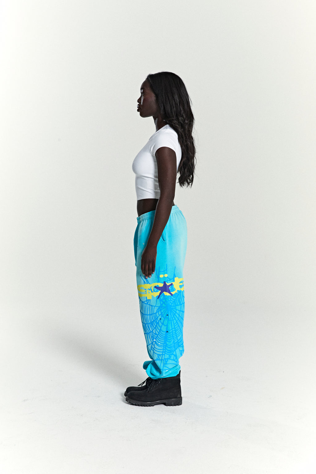 Woman in blue HEAVY WEB SWEATPANTS with SP5DER web graphic, scattered stars, and elasticized cuffs.