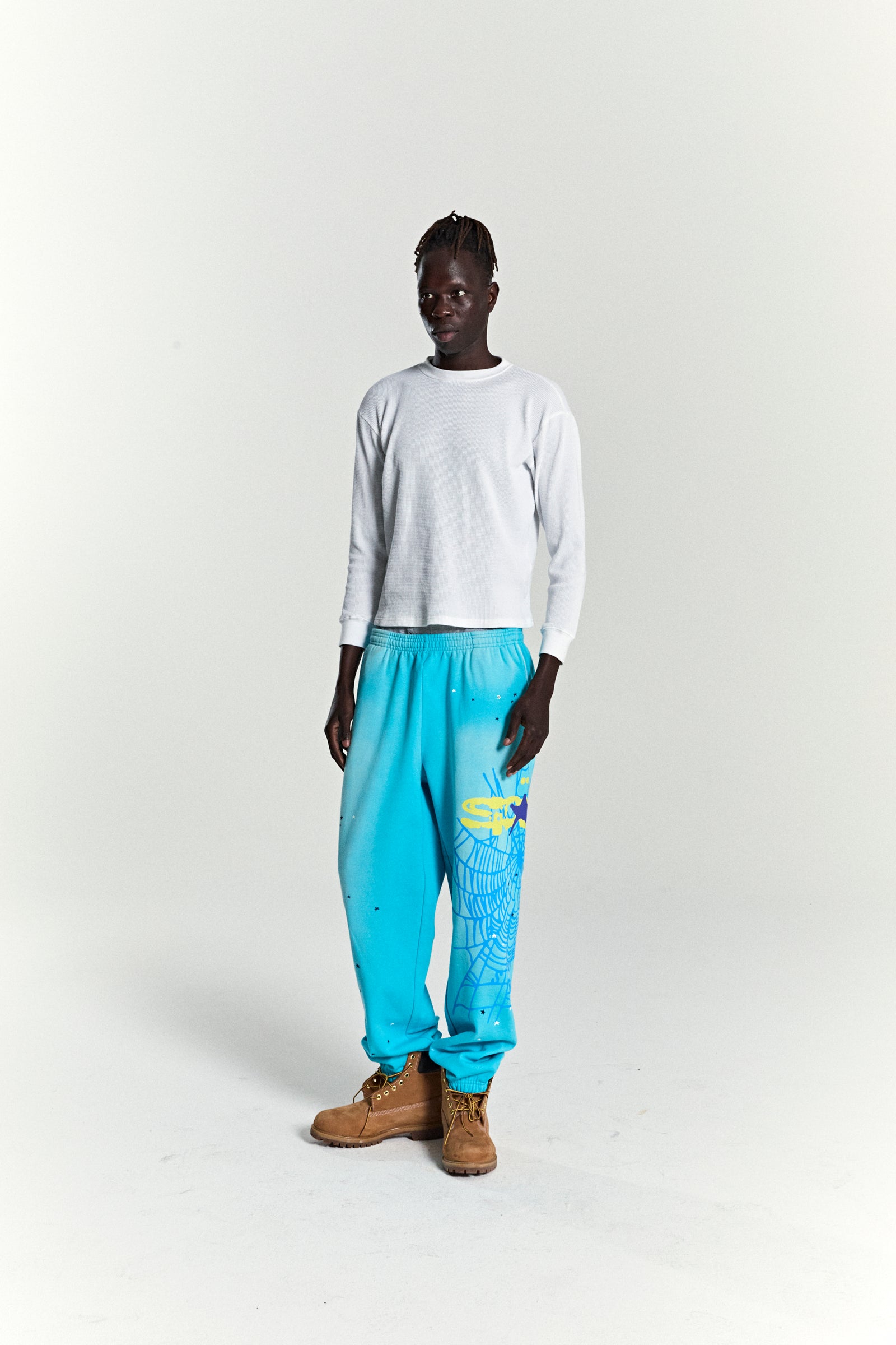 Model wearing turquoise HEAVY WEB SWEATPANTS with SP5DER web graphic, scattered stars, and elasticized cuffs.