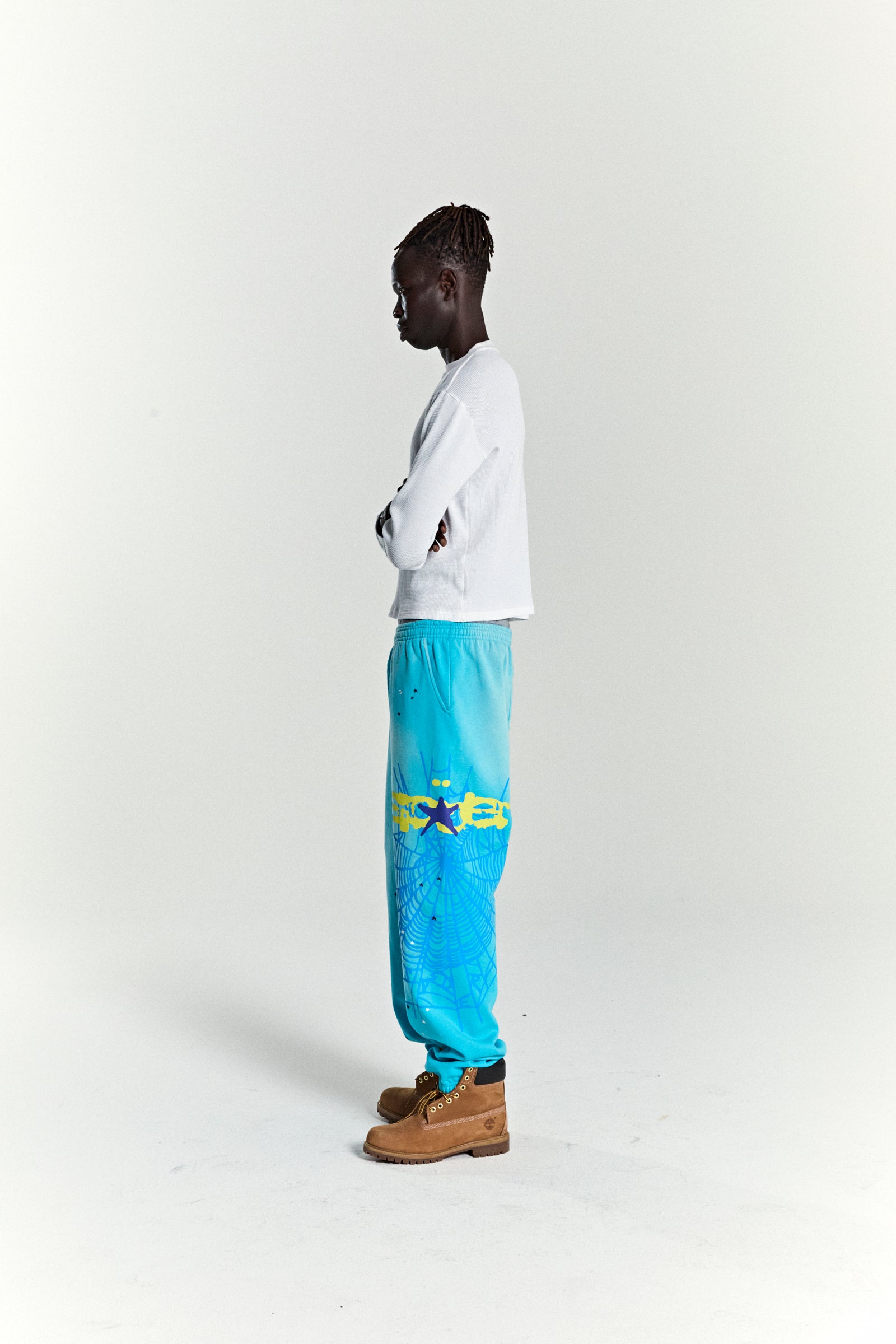 Model in turquoise HEAVY WEB SWEATPANTS with yellow SP5DER web graphic and scattered stars.