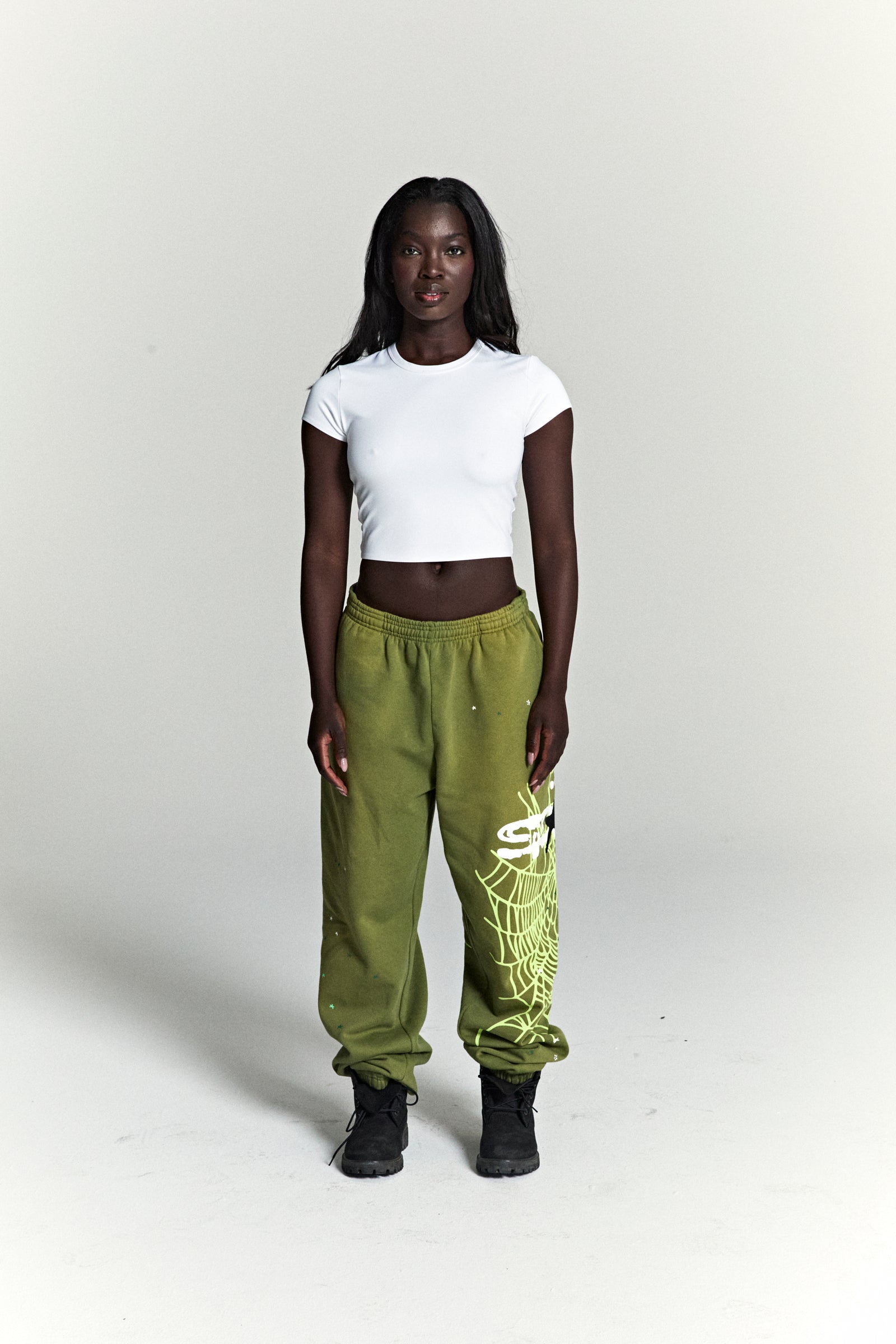 Woman in olive green HEAVY WEB SWEATPANTS with SP5DER web graphic and scattered star print.