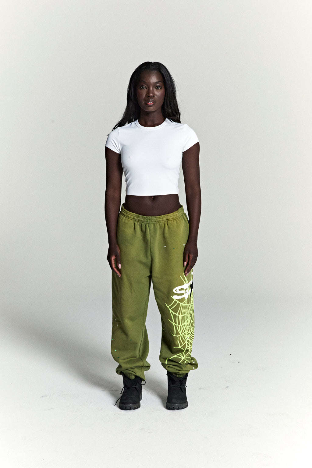 Woman in olive green HEAVY WEB SWEATPANTS with SP5DER web graphic and scattered star print.