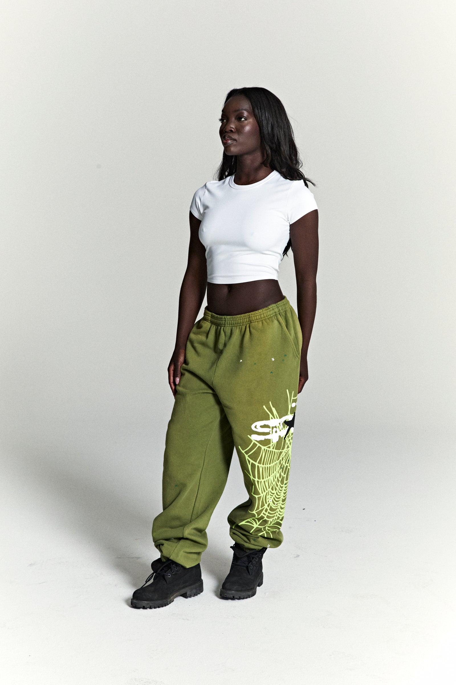 Model wears olive green HEAVY WEB SWEATPANTS with SP5DER web graphic, scattered stars, and elasticized cuffs.