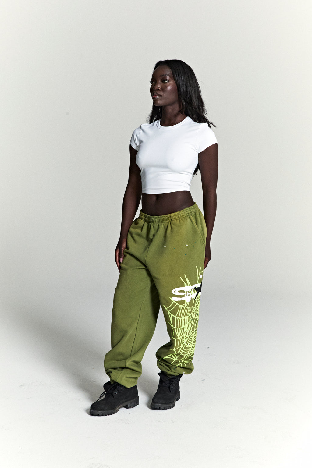 Model wears olive green HEAVY WEB SWEATPANTS with SP5DER web graphic, scattered stars, and elasticized cuffs.