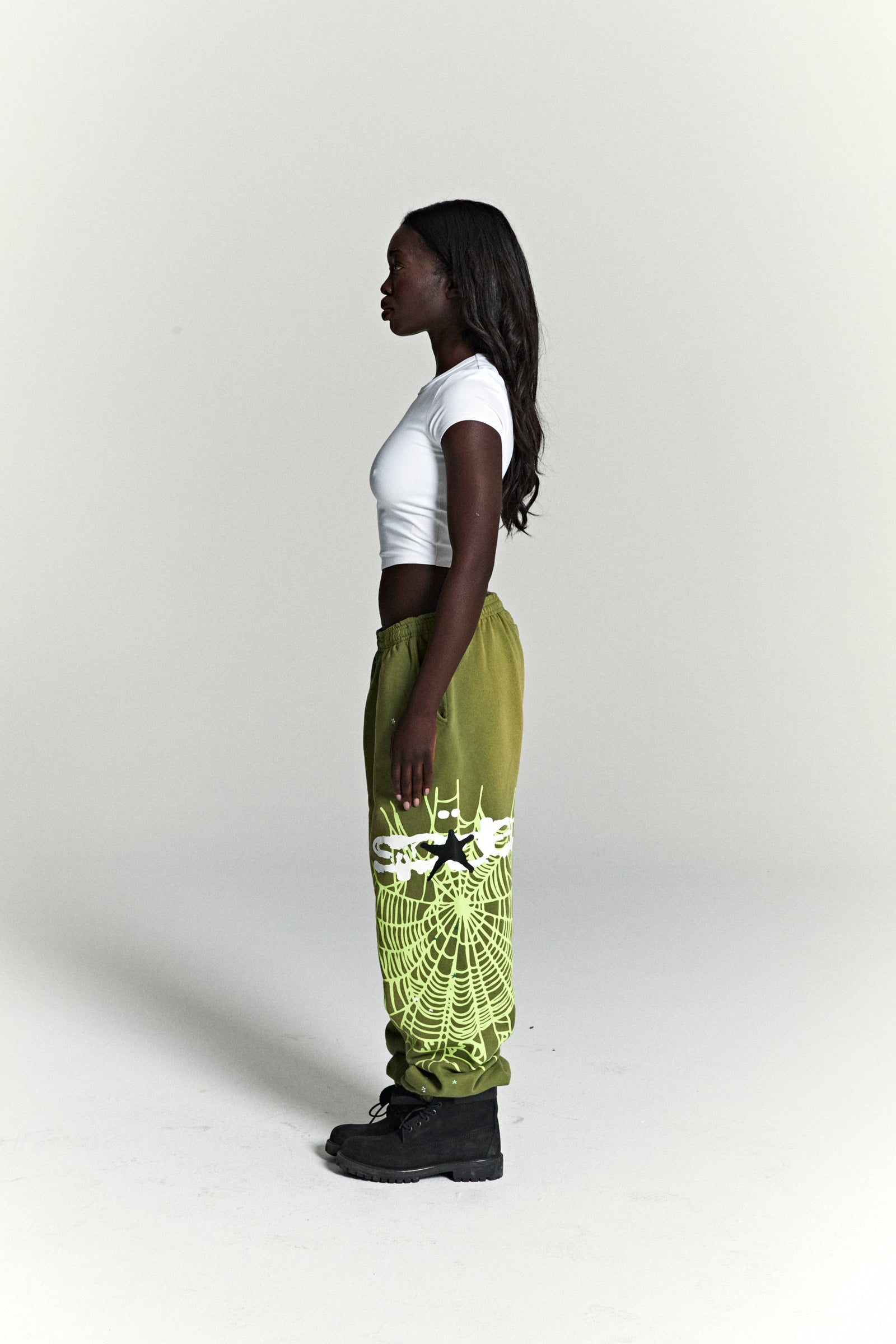Woman in olive green HEAVY WEB SWEATPANTS with neon green SP5DER web graphic and scattered stars.