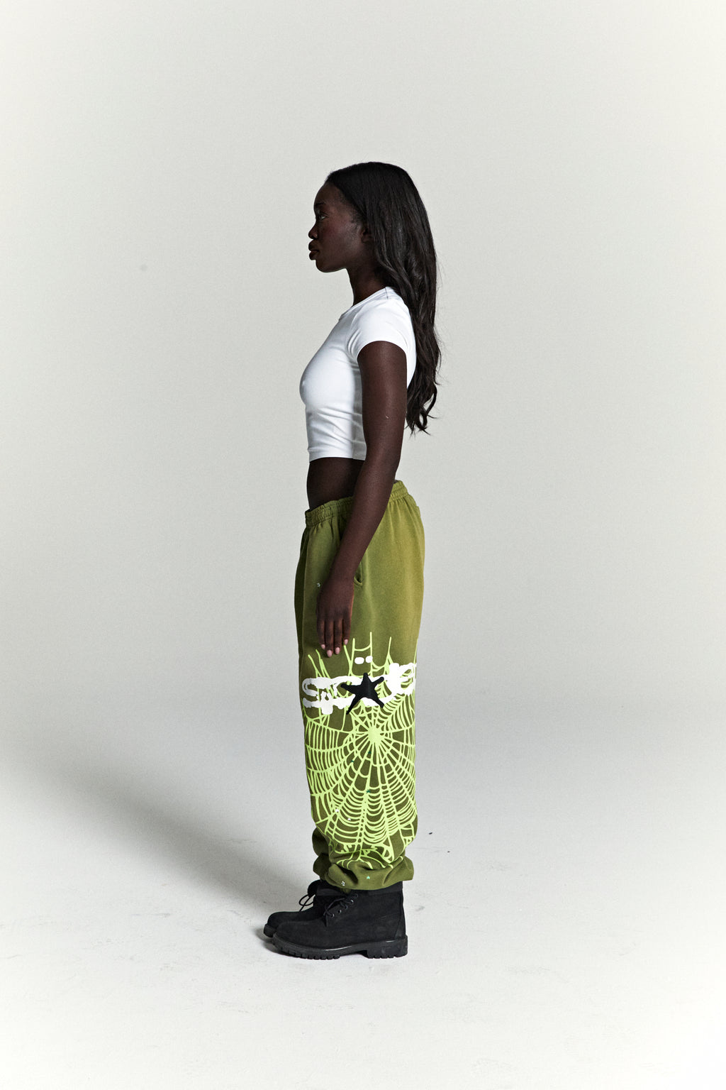 Woman in olive green HEAVY WEB SWEATPANTS with neon green SP5DER web graphic and scattered stars.