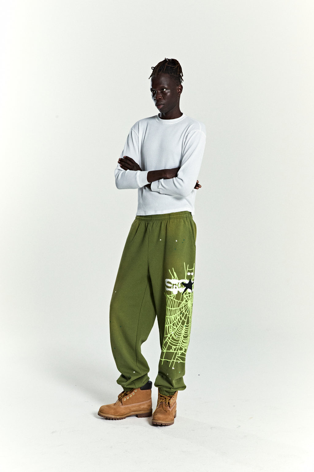 Model wearing olive green HEAVY WEB SWEATPANTS with SP5DER web graphic and scattered star print.
