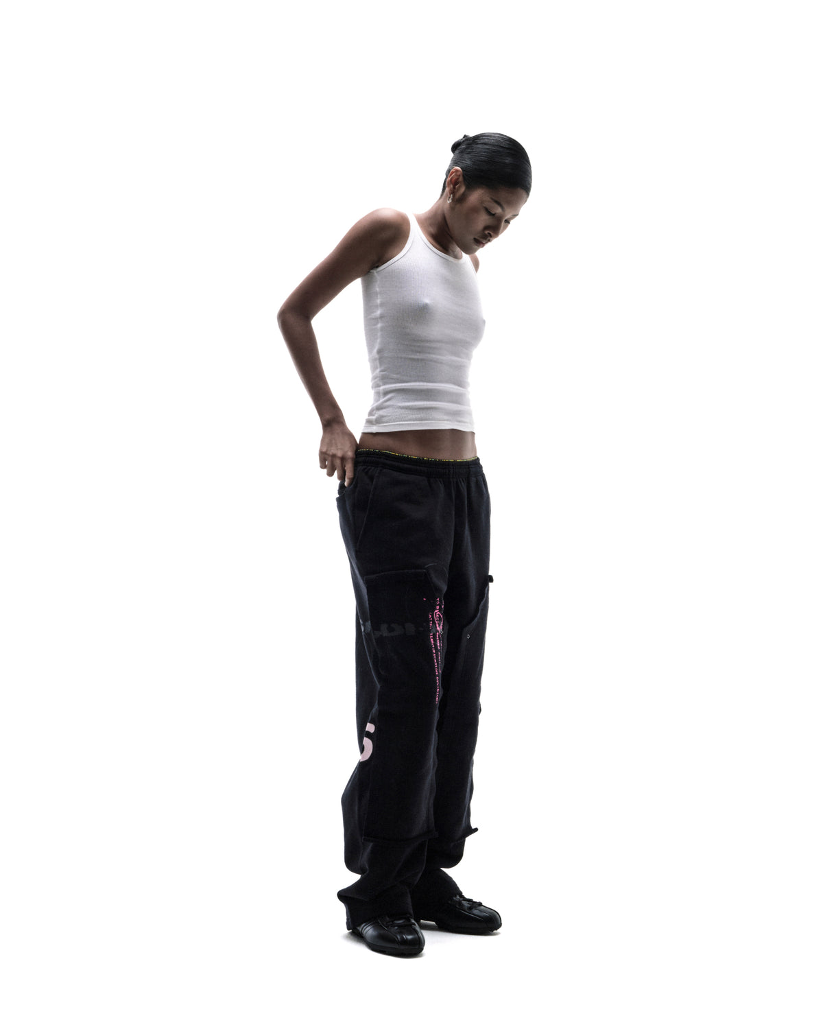 A model wearing a white ribbed tank top and black baggy cargo pants with pink graphic details against a white background.
