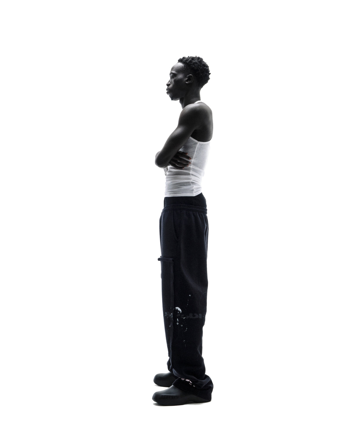 A model in profile wearing a white ribbed tank top, black cargo sweatpants, and dark slip-on shoes against a white background.