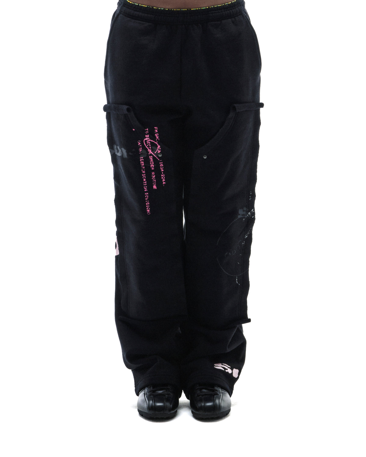 Model wearing black fleece cargo sweatpants with pink graphic text and layered pocket detailing on a white background.