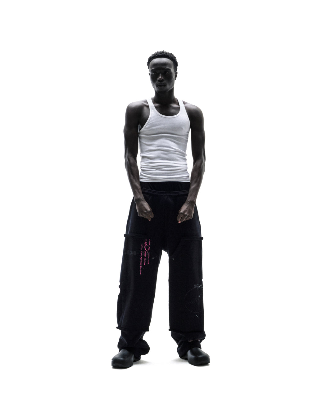 A model wearing a white ribbed tank top and black oversized cargo sweatpants with pink graphic details against a white background.