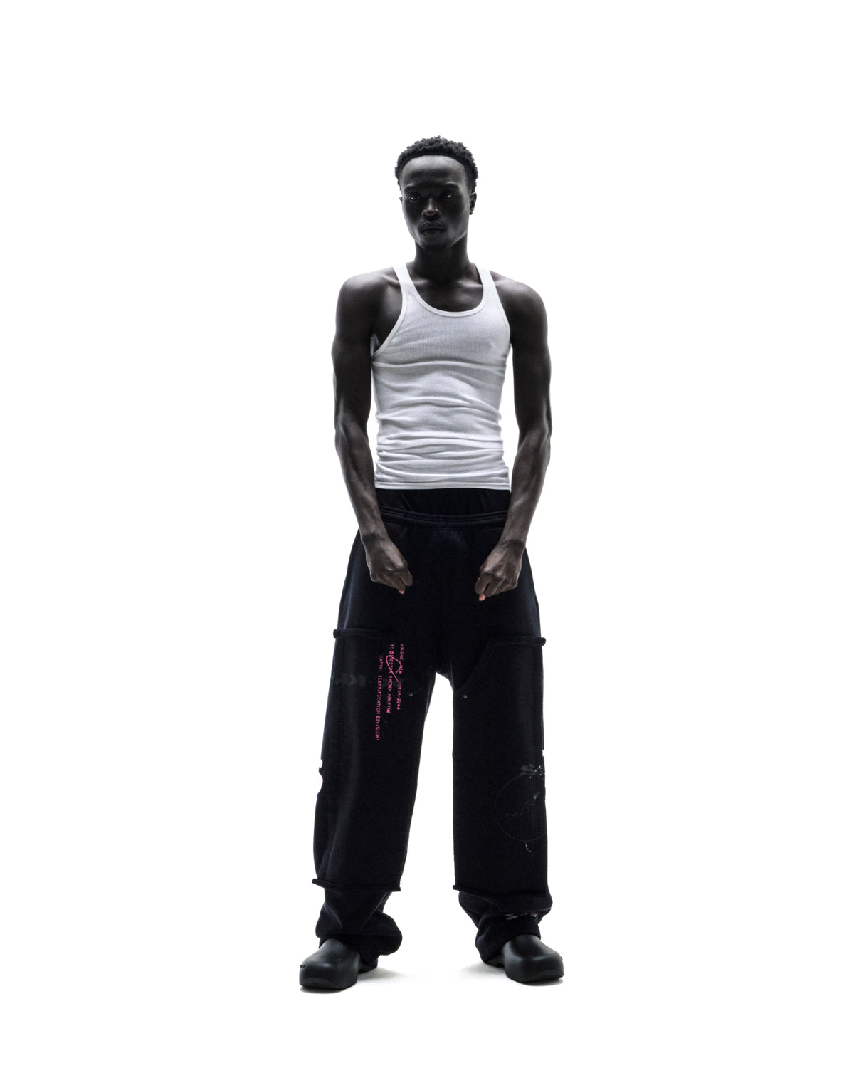 A model wearing a white ribbed tank top and black oversized cargo sweatpants with pink graphic details against a white background.