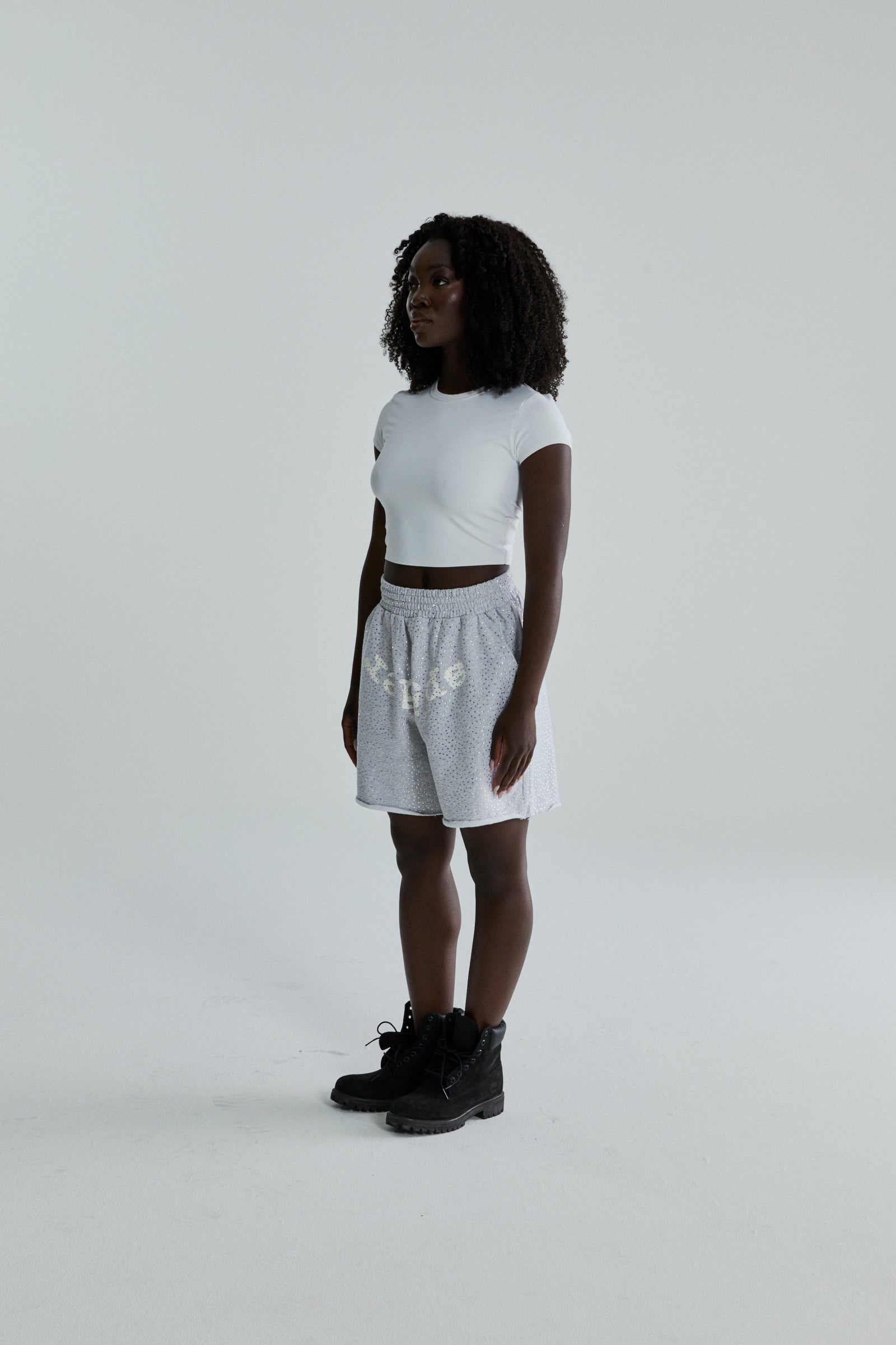 Woman wearing gray VVS SWEATSHORT with tonal rhinestones, SP5DER text, and raw hem.