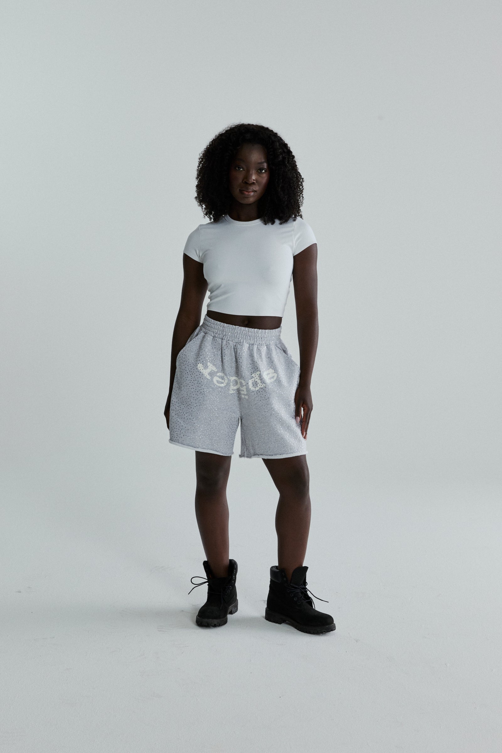Woman wearing grey VVS SWEATSHORT with tonal rhinestones, SP5DER text, and raw hem.