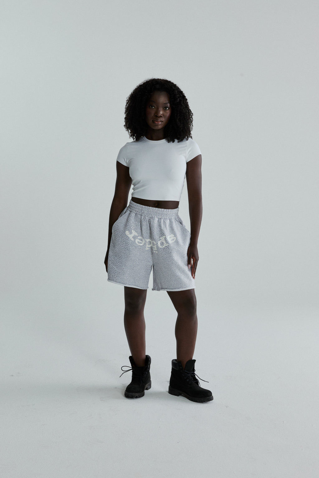 Woman wearing grey VVS SWEATSHORT with tonal rhinestones, SP5DER text, and raw hem.