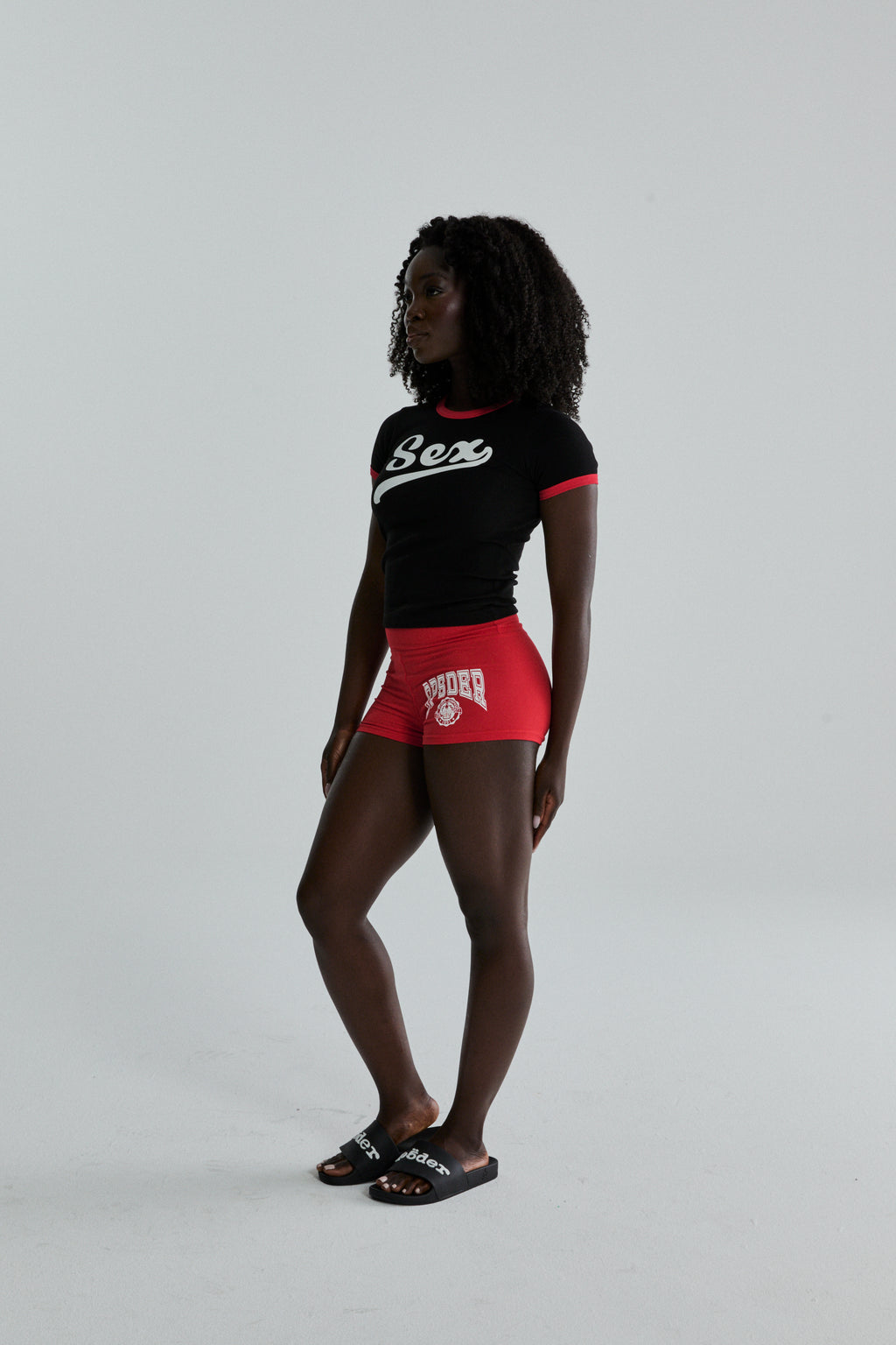 Black cropped cotton/spandex tee with "Sex" graphic, red ribbed trim, paired with red shorts and black slides.