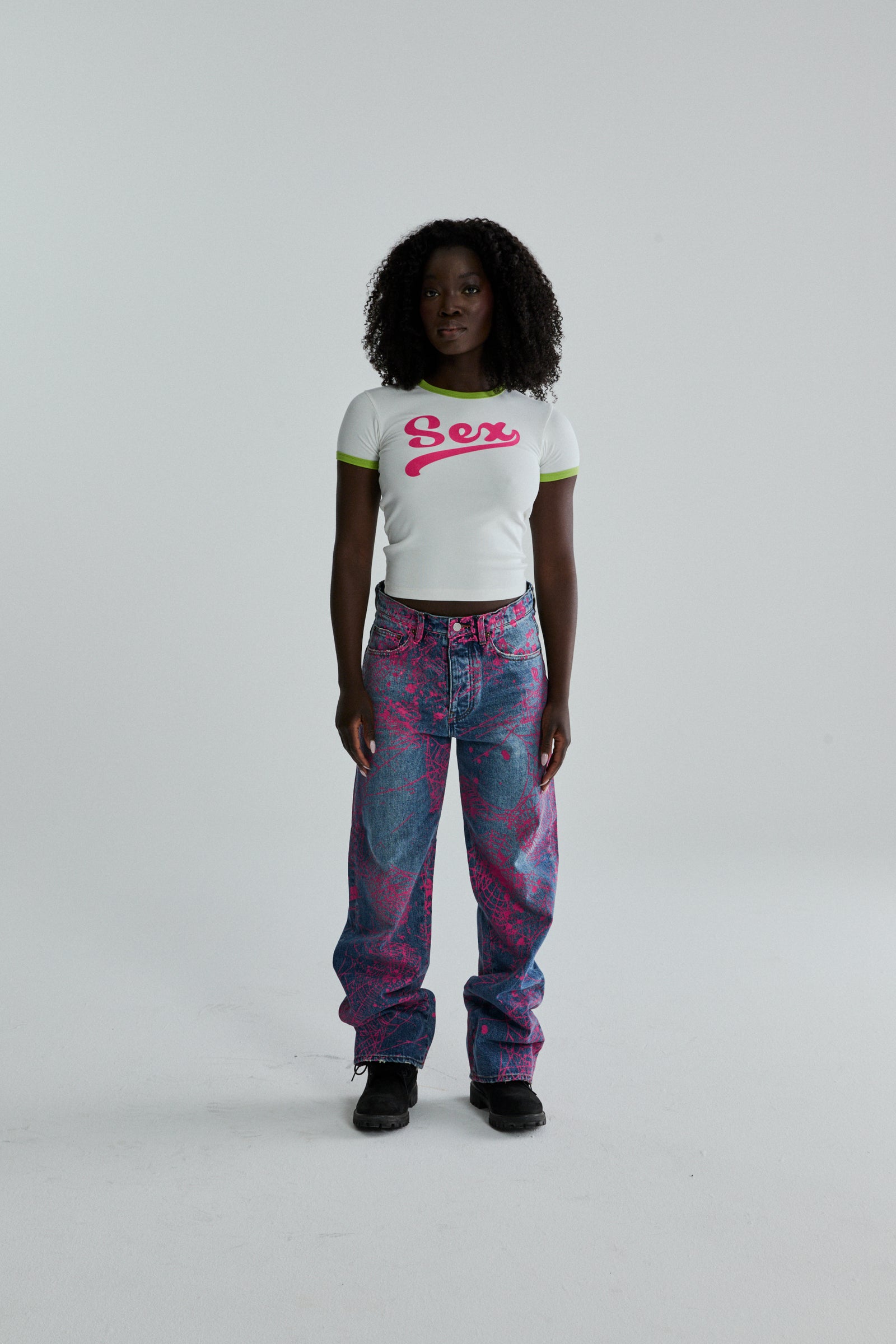 White cropped cotton/spandex tee with pink "Sex" graphic and green ribbed trim, paired with patterned jeans.
