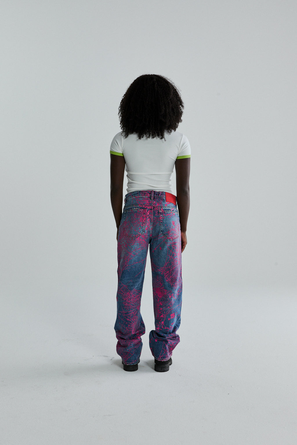 Woman from behind wearing a white cropped tee with green trim and baggy pink and blue graphic print jeans.