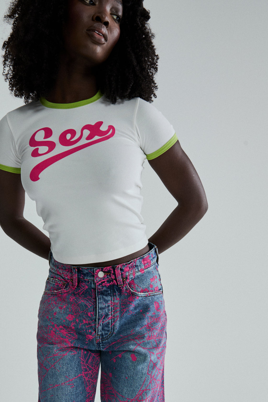 White cropped cotton/spandex tee with "Sex" graphic in pink, green ribbed trim, paired with blue patterned jeans.