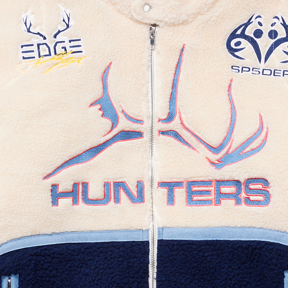 Cream sherpa moto jacket with blue "HUNTERS" embroidery, antler designs, SP5DER patches, and a silver zip closure.