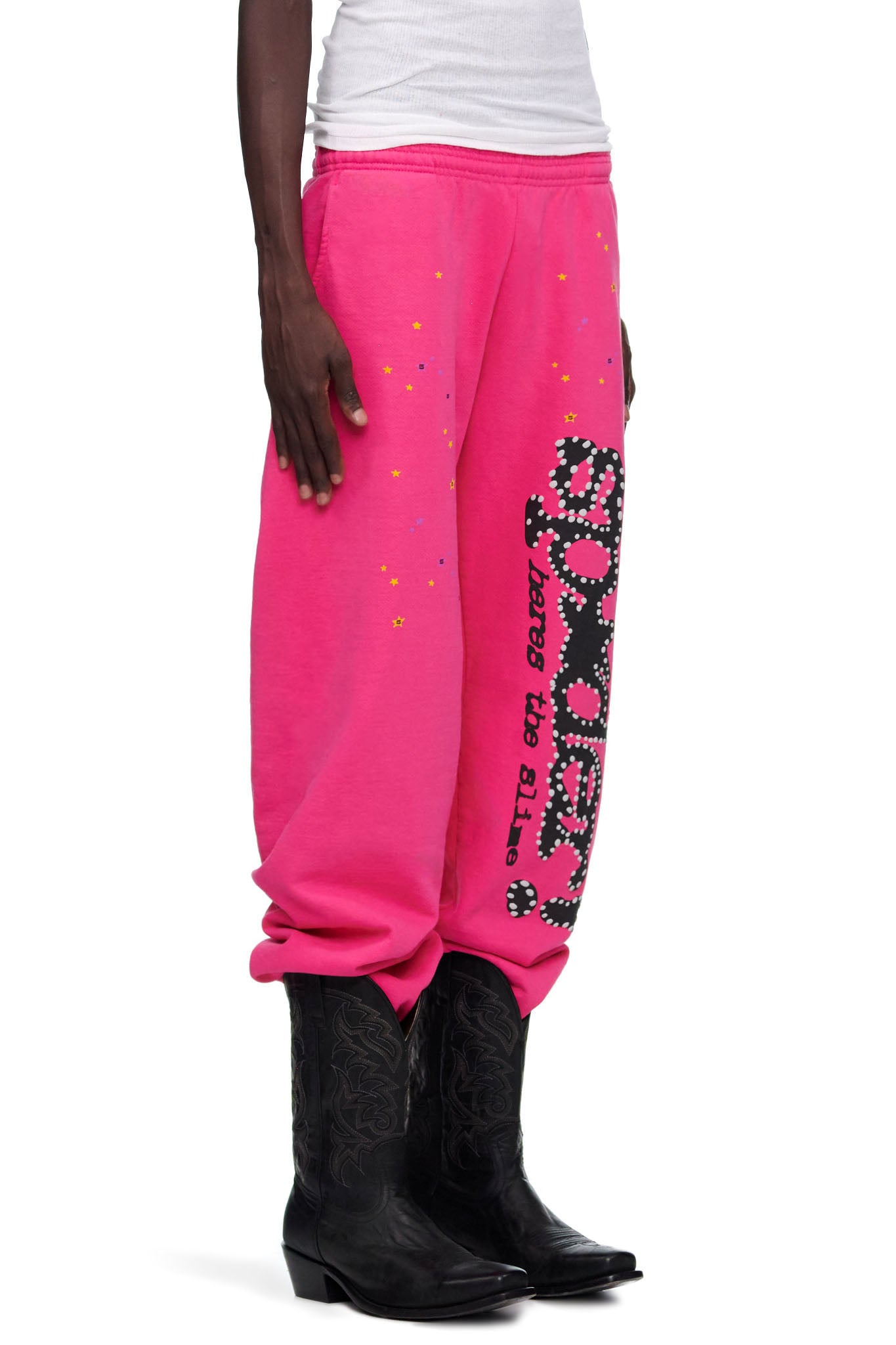 Pink P*NK V2 sweatpants with SP5DER motif, star prints, and "heres the slime" text, worn with black cowboy boots.