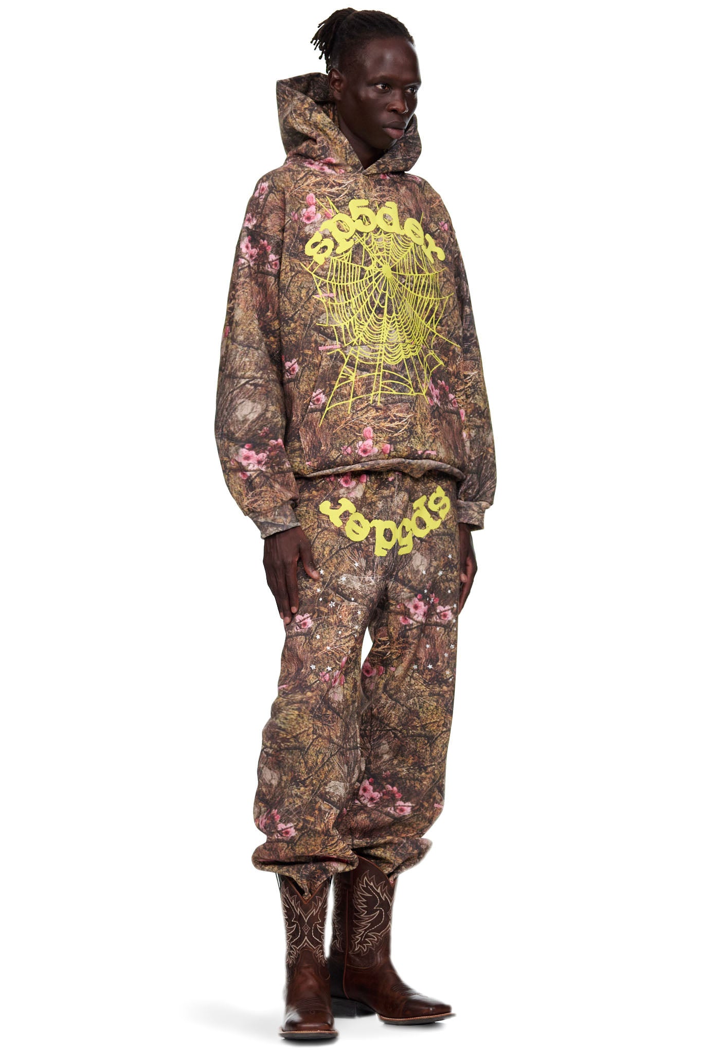 Male model in REAL TREE OG WEB SWEATPANT, brown camo with pink flowers, yellow puff print spiderweb and text.