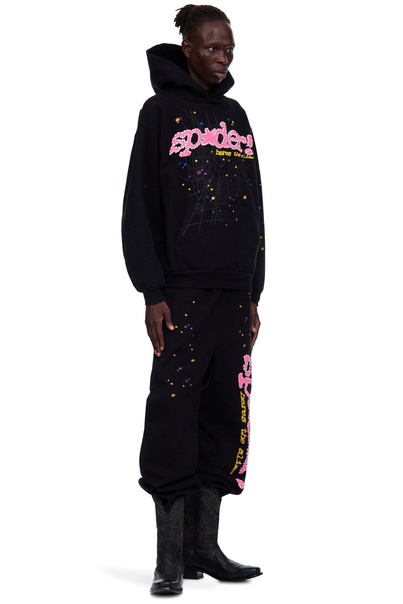 Male model in black SP5DER hoodie and sweatpants with pink puff print, spiderweb, and star designs.