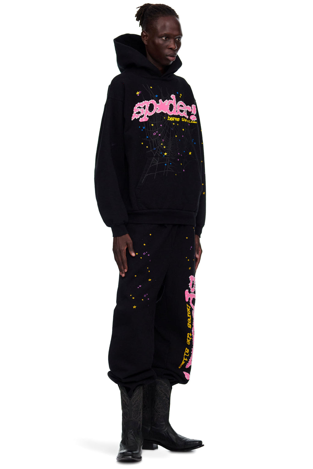 Male model in black SP5DER hoodie and sweatpants with pink puff print, spiderweb, and star designs.