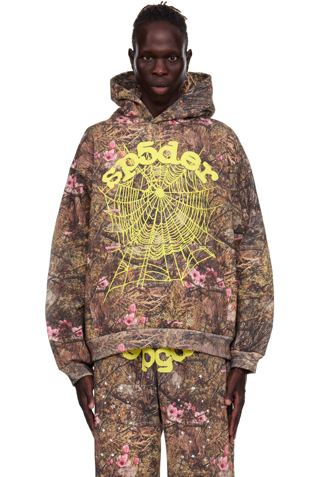 Male model in Real Tree OG Web Hoodie, a brown camo fleece with pink flowers, yellow "Sp5der" text, and a large spider web print.