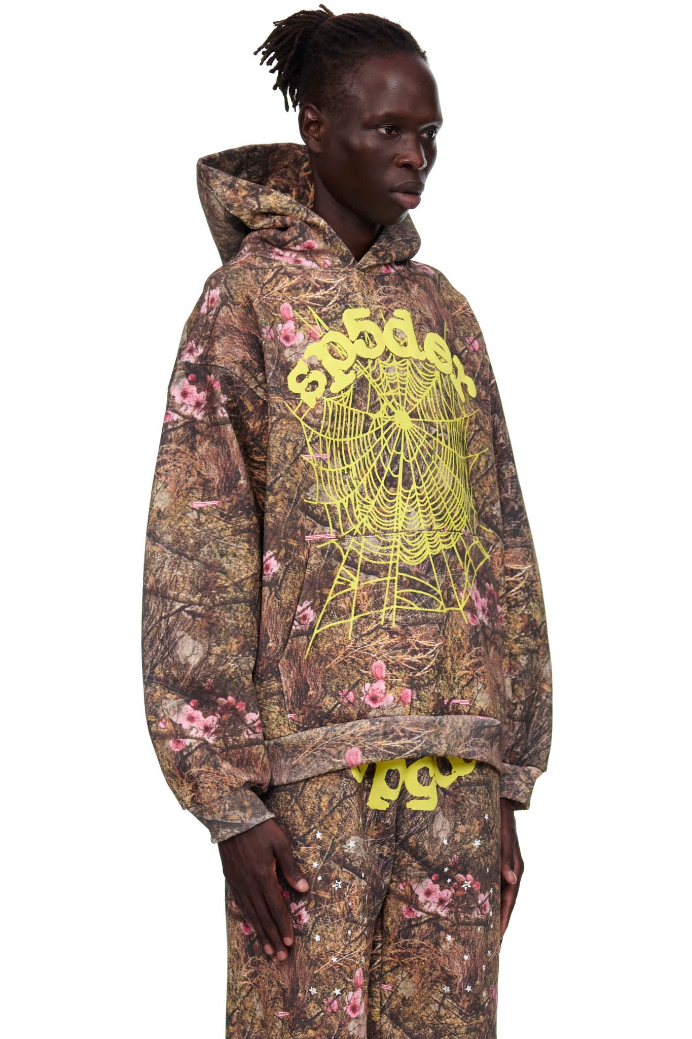 Male model in Real Tree OG Web Hoodie, brown camo with pink flowers, yellow "SP5DER" text and web print.