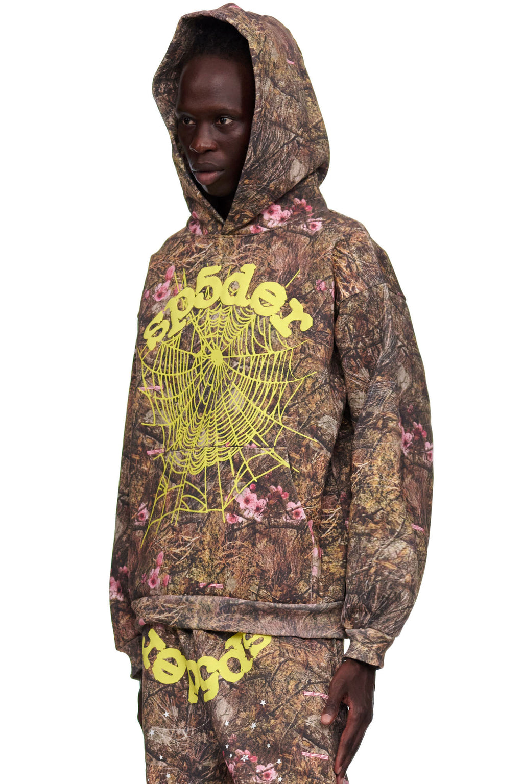 Male model in a Real Tree OG Web Hoodie, featuring brown camo with pink flowers and a yellow puff print spider web and text.