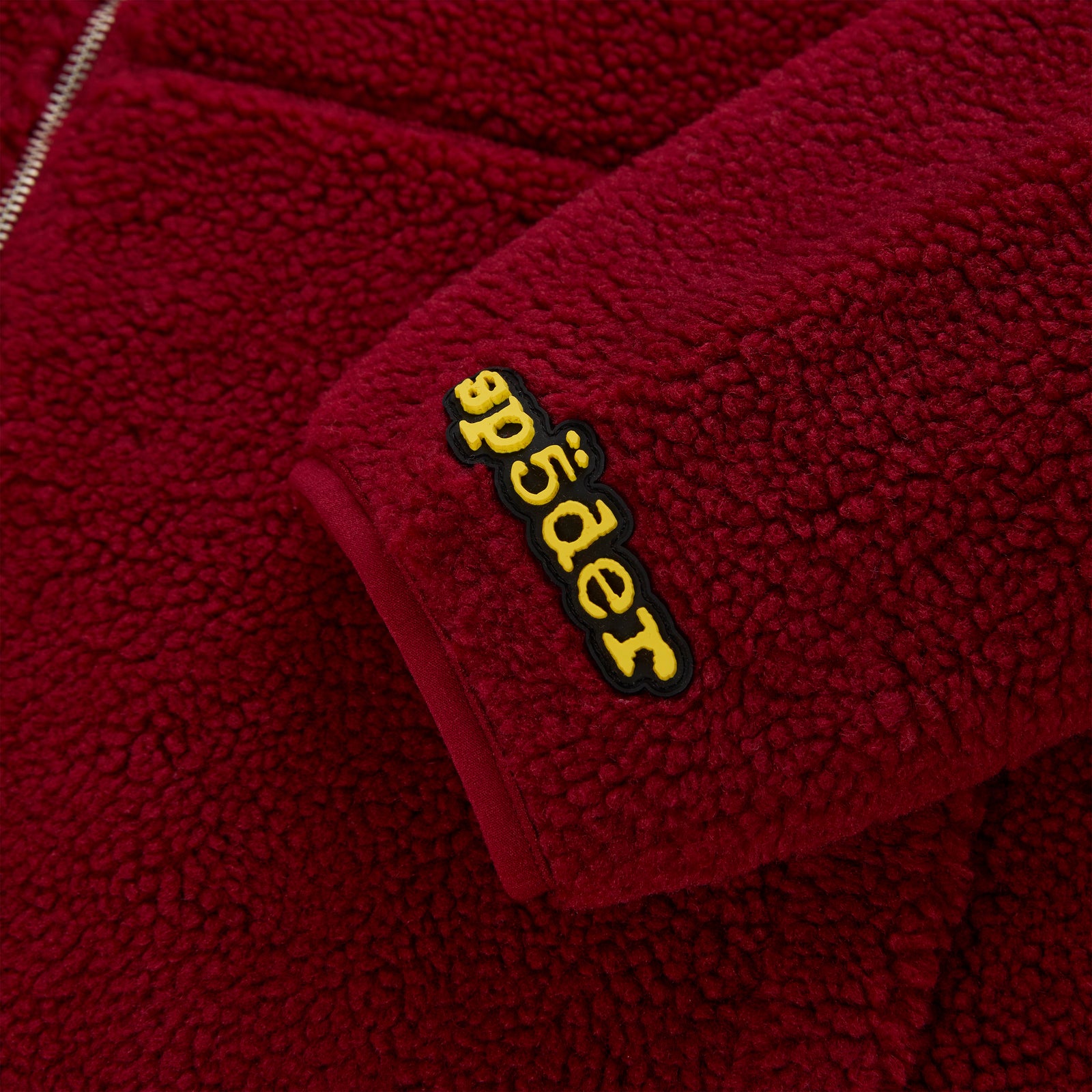 Close-up of a red sherpa fleece zip-up jacket sleeve with a yellow rubberized "Spyder" logo patch.