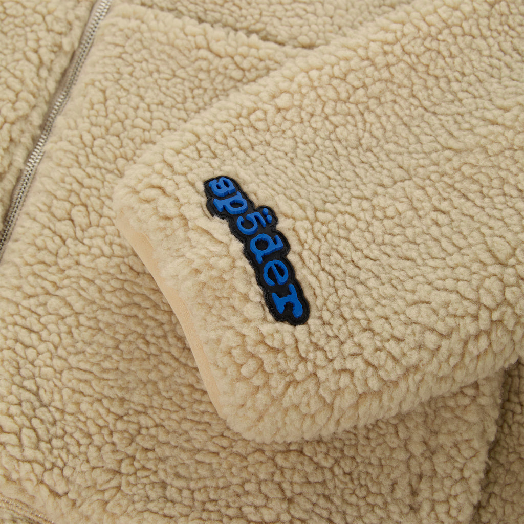 Close-up of a beige teddy sherpa fleece zip-up jacket with a blue rubberized logo patch on the wrist.