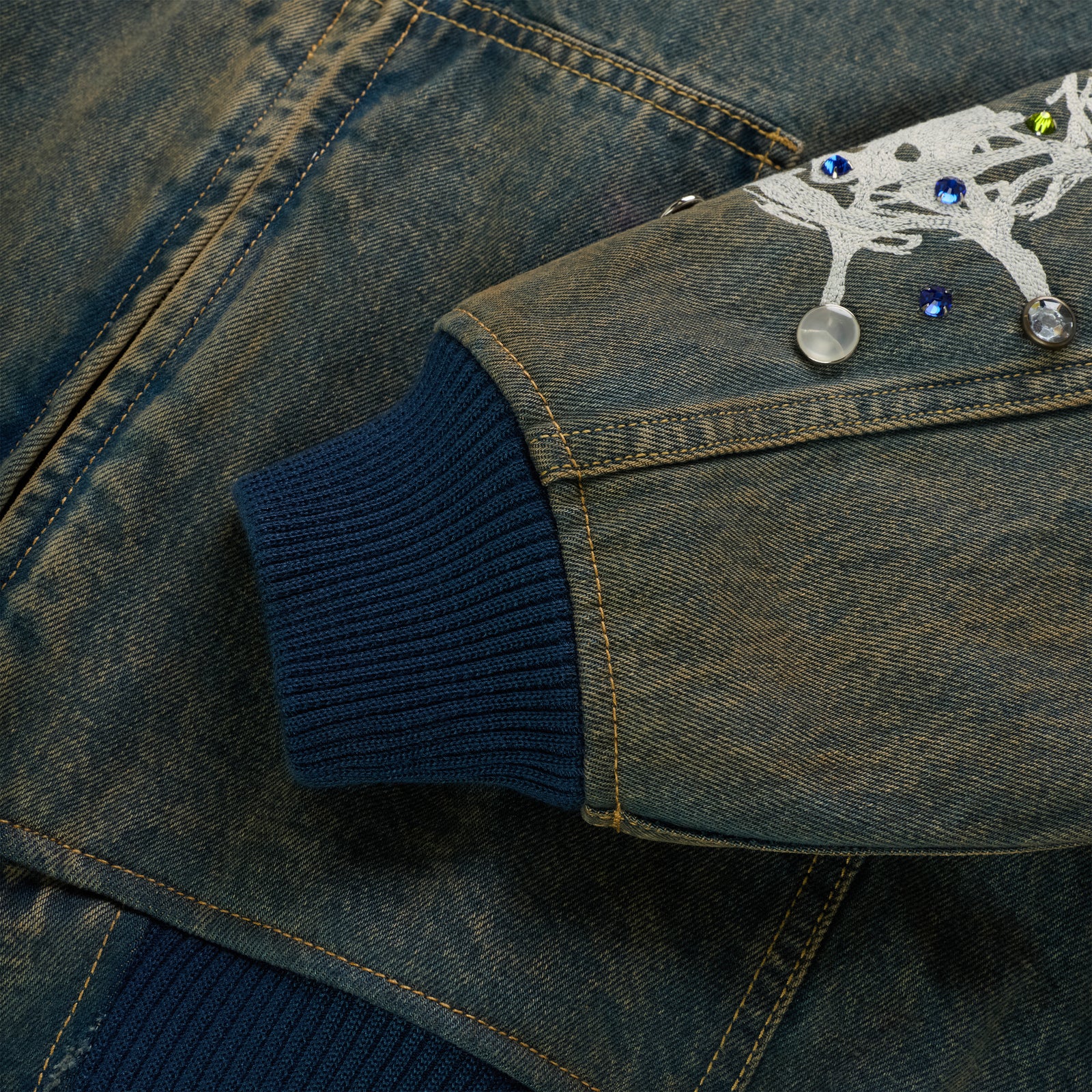 Faded denim jacket sleeve with blue rib-knit cuff, white web print, and rhinestone embellishments.