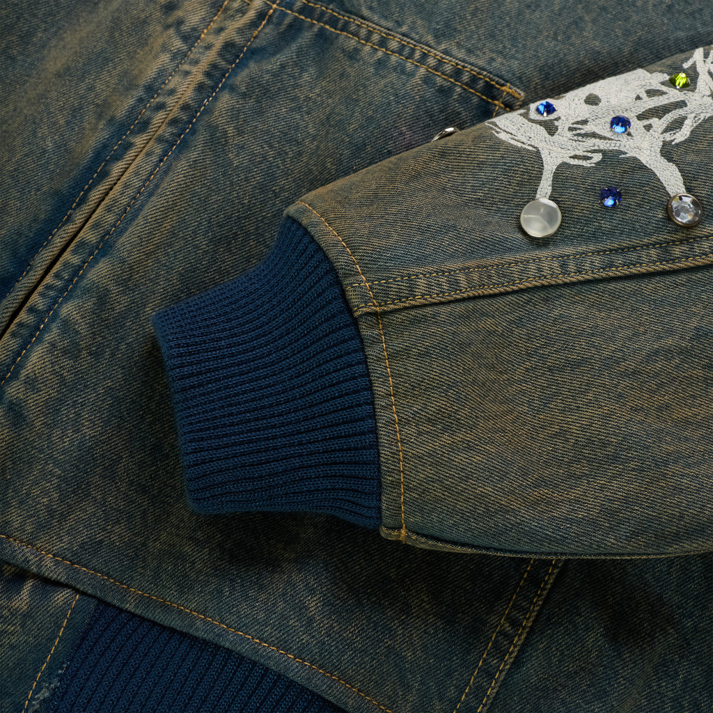 Faded denim jacket sleeve with blue rib-knit cuff, white web print, and rhinestone embellishments.