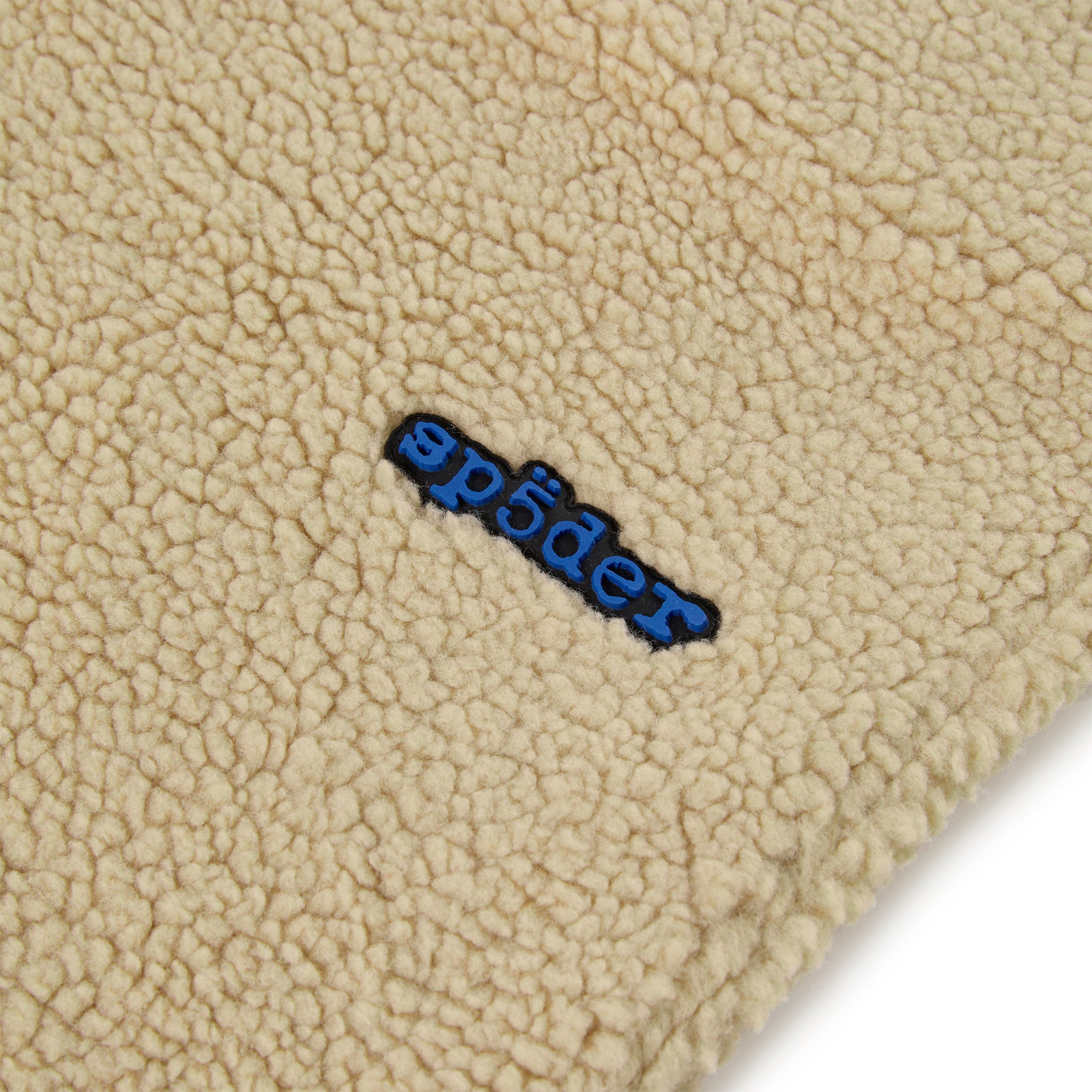 Close-up of a cream teddy fleece pant with a blue rubberized "sp5der" logo patch.
