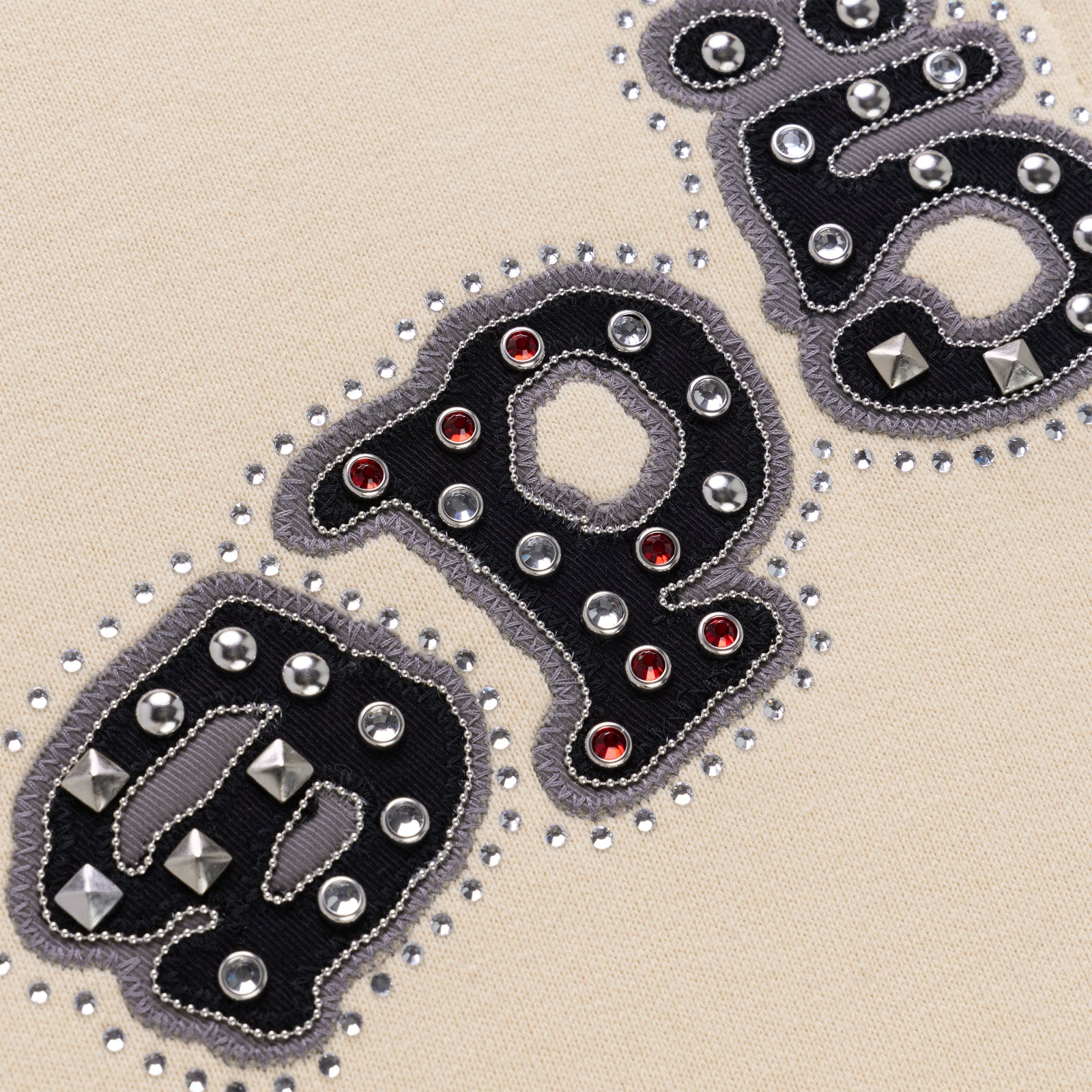 Close-up of "OG" logo on beige cotton fleece, embellished with mixed metal studs, dome rivets, and red pyramid spikes.