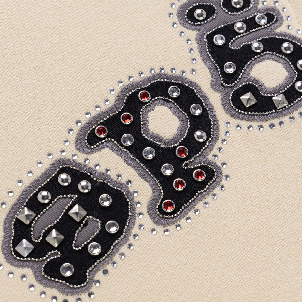 Close-up of "OG" logo on beige cotton fleece, embellished with mixed metal studs, dome rivets, and red pyramid spikes.