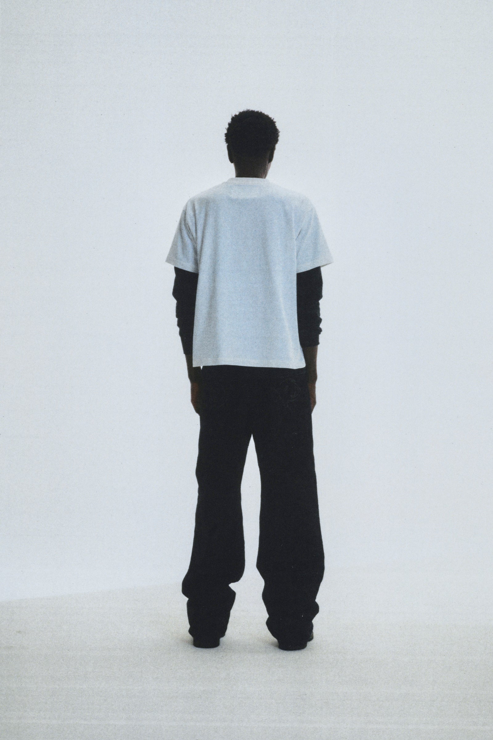 Man from behind wearing a light grey 100% cotton COMMERCE TEE over a black long-sleeve shirt and black pants.