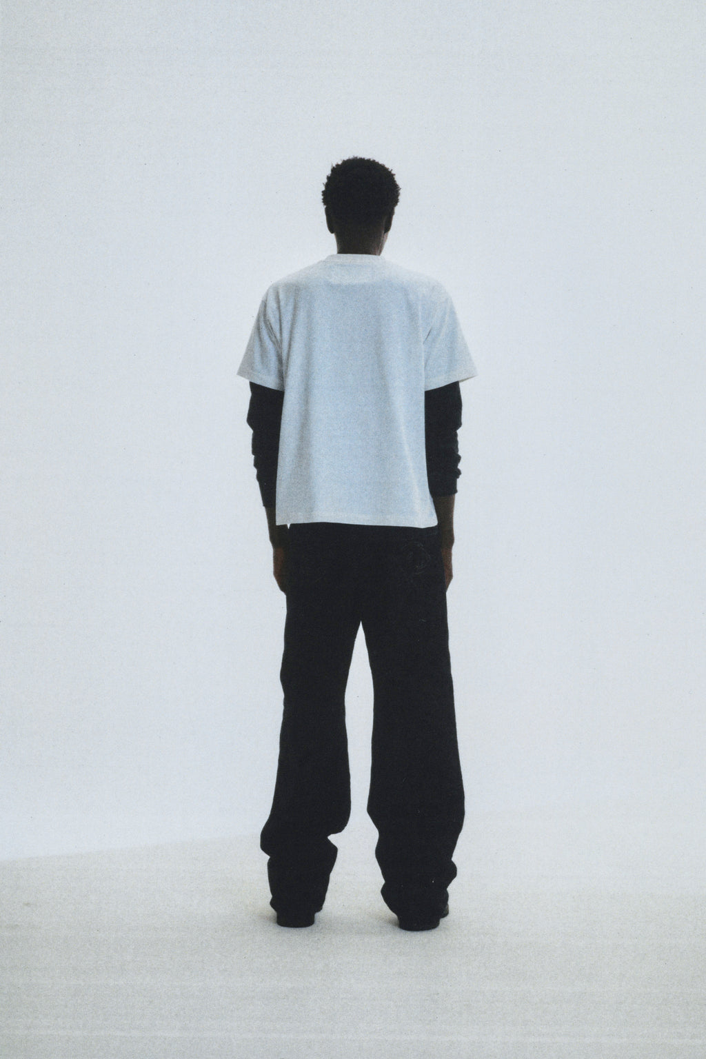 Man from behind wearing a light grey 100% cotton COMMERCE TEE over a black long-sleeve shirt and black pants.