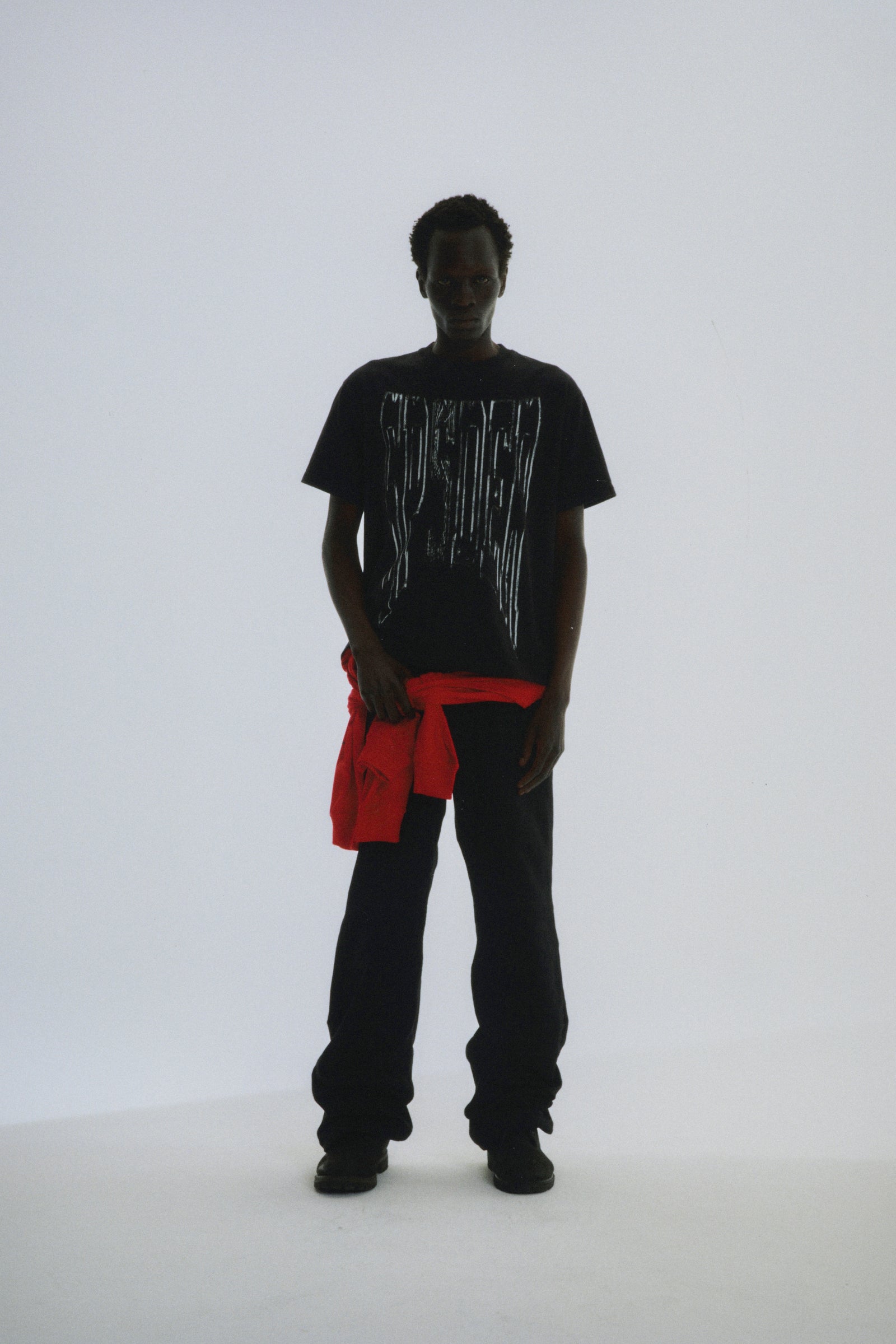 Black DIFFUSED COLLEGIATE TEE with white graphic print, worn by a model with a red garment tied around the waist.