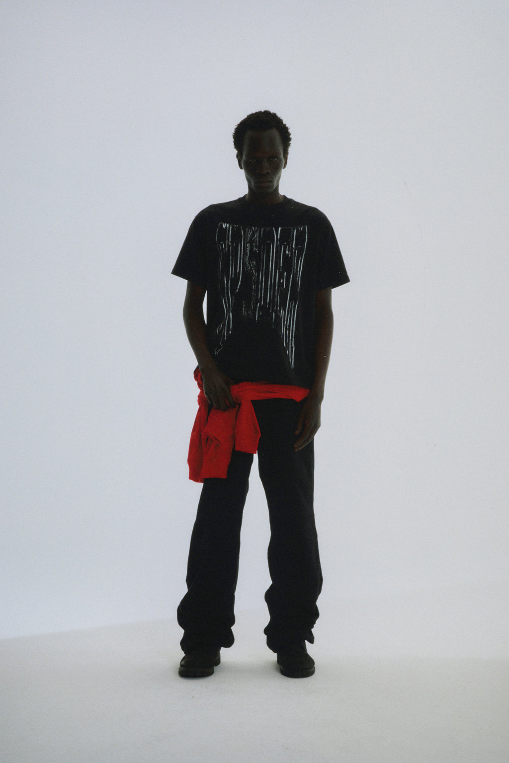 Black DIFFUSED COLLEGIATE TEE with white graphic print, worn by a model with a red garment tied around the waist.
