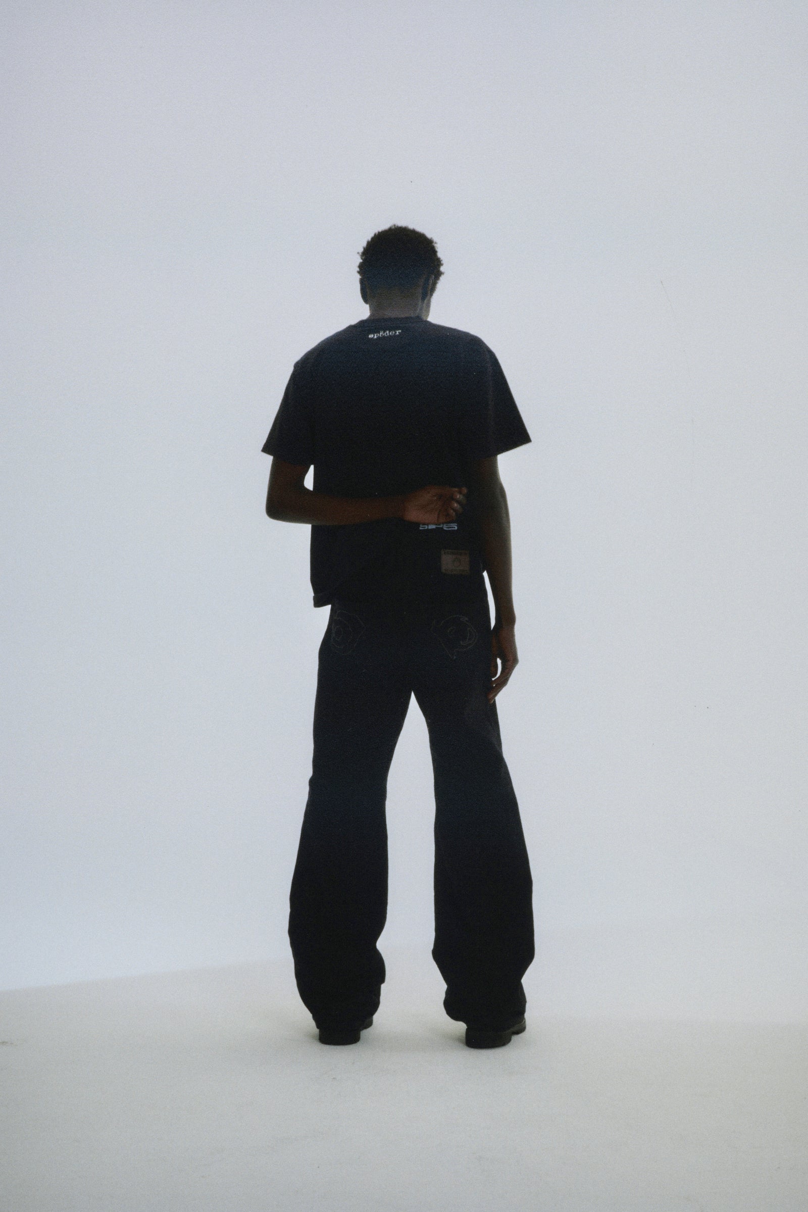 Man in black DIFFUSED COLLEGIATE TEE with 'SP5DER' logo on back neck, paired with black pants.