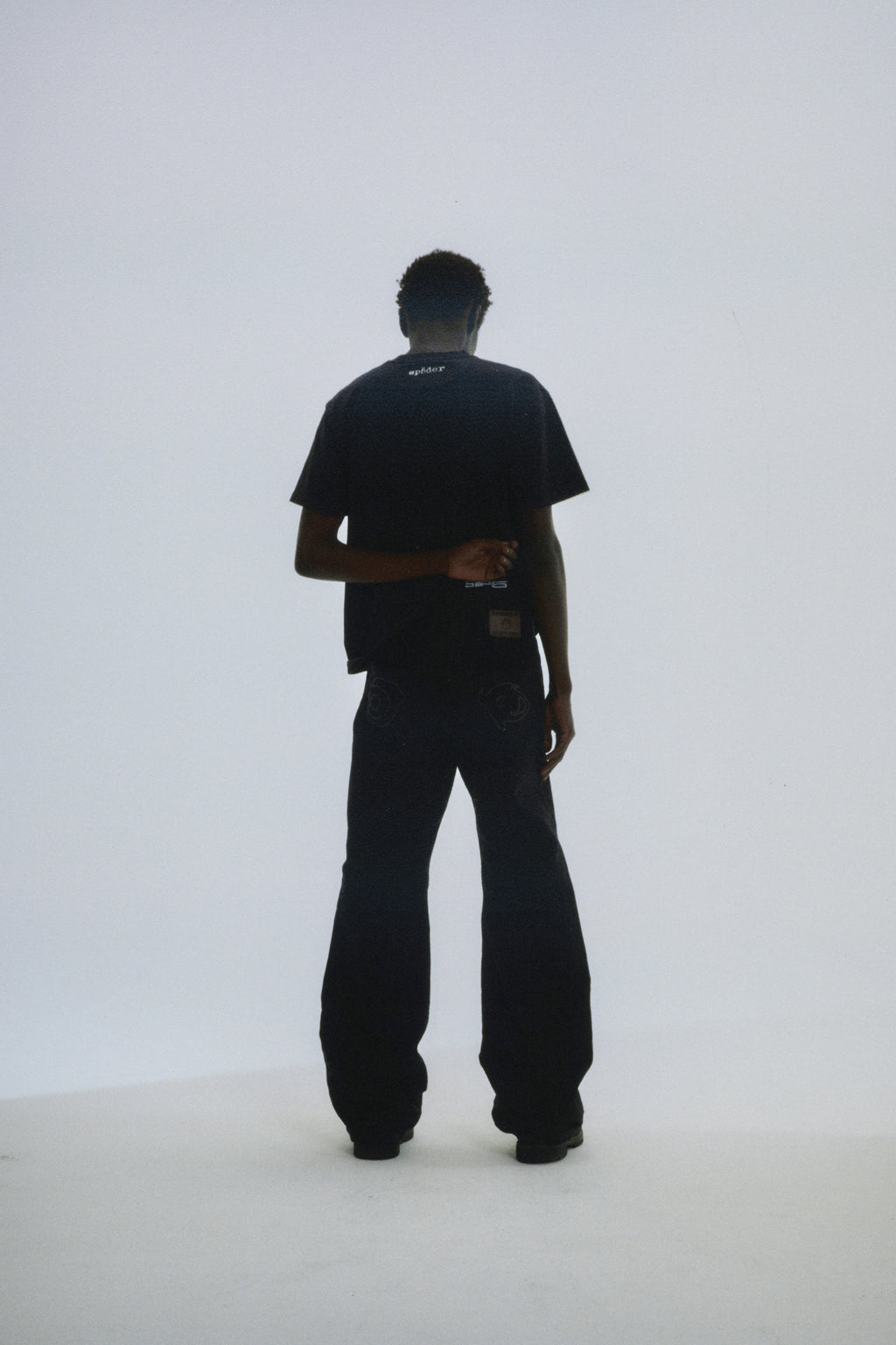 Man in black DIFFUSED COLLEGIATE TEE with 'SP5DER' logo on back neck, paired with black pants.