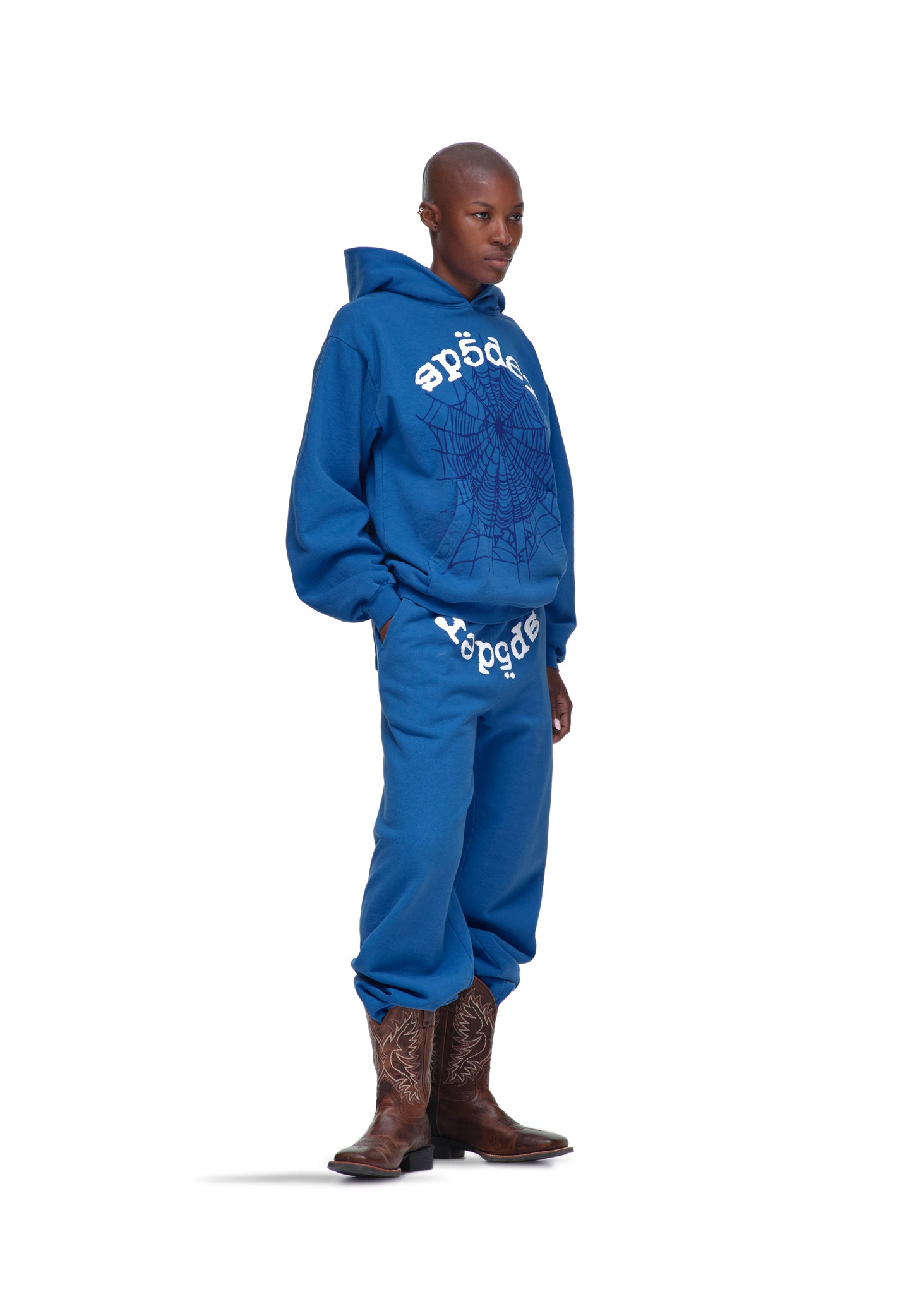Male model in blue Legacy Hoodie and sweatpants with white spider web and text print, wearing cowboy boots.