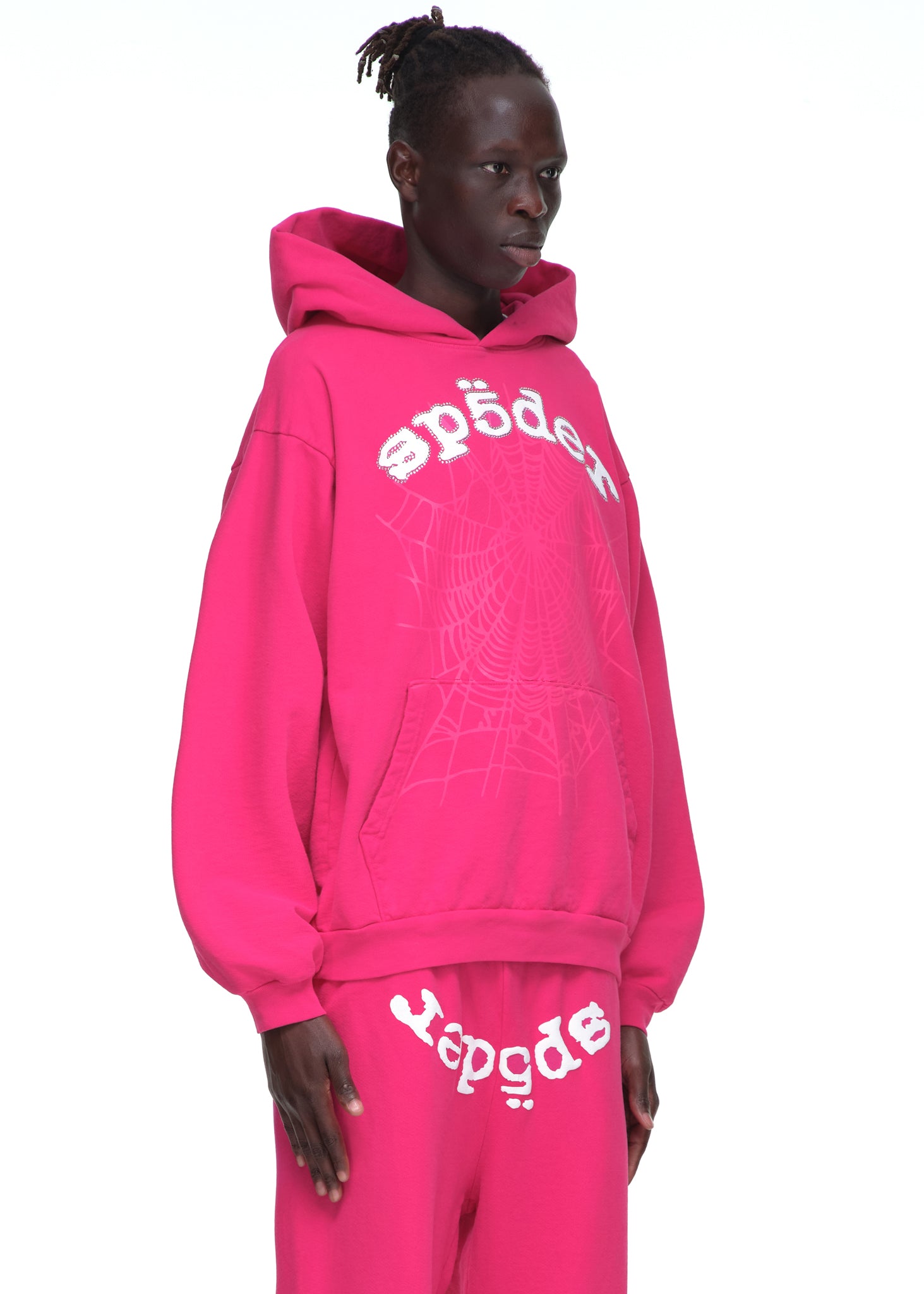 Male model in a pink LEGACY HOODIE with rhinestone SP5DER text and a web print, paired with matching sweatpants.