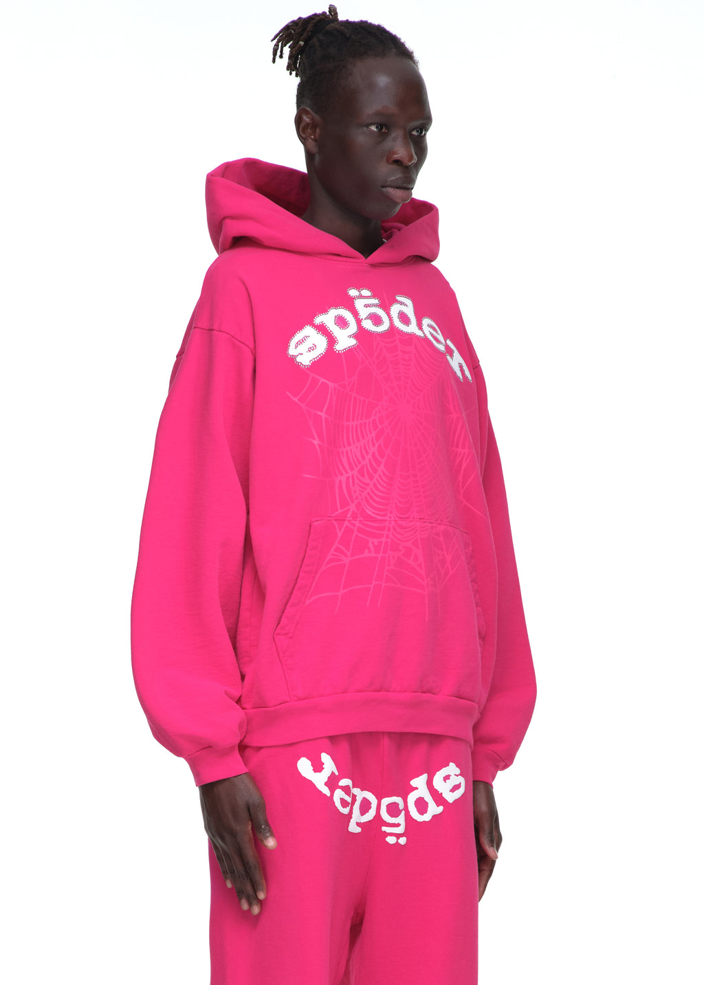 Male model in a pink LEGACY HOODIE with rhinestone SP5DER text and a web print, paired with matching sweatpants.