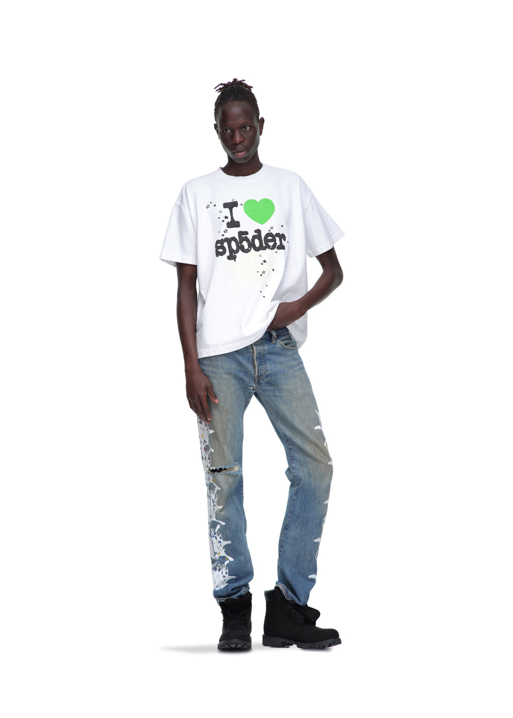 Male model in a white 14oz cotton Souvenir Tee with green heart graphic, distressed blue jeans, and black boots.
