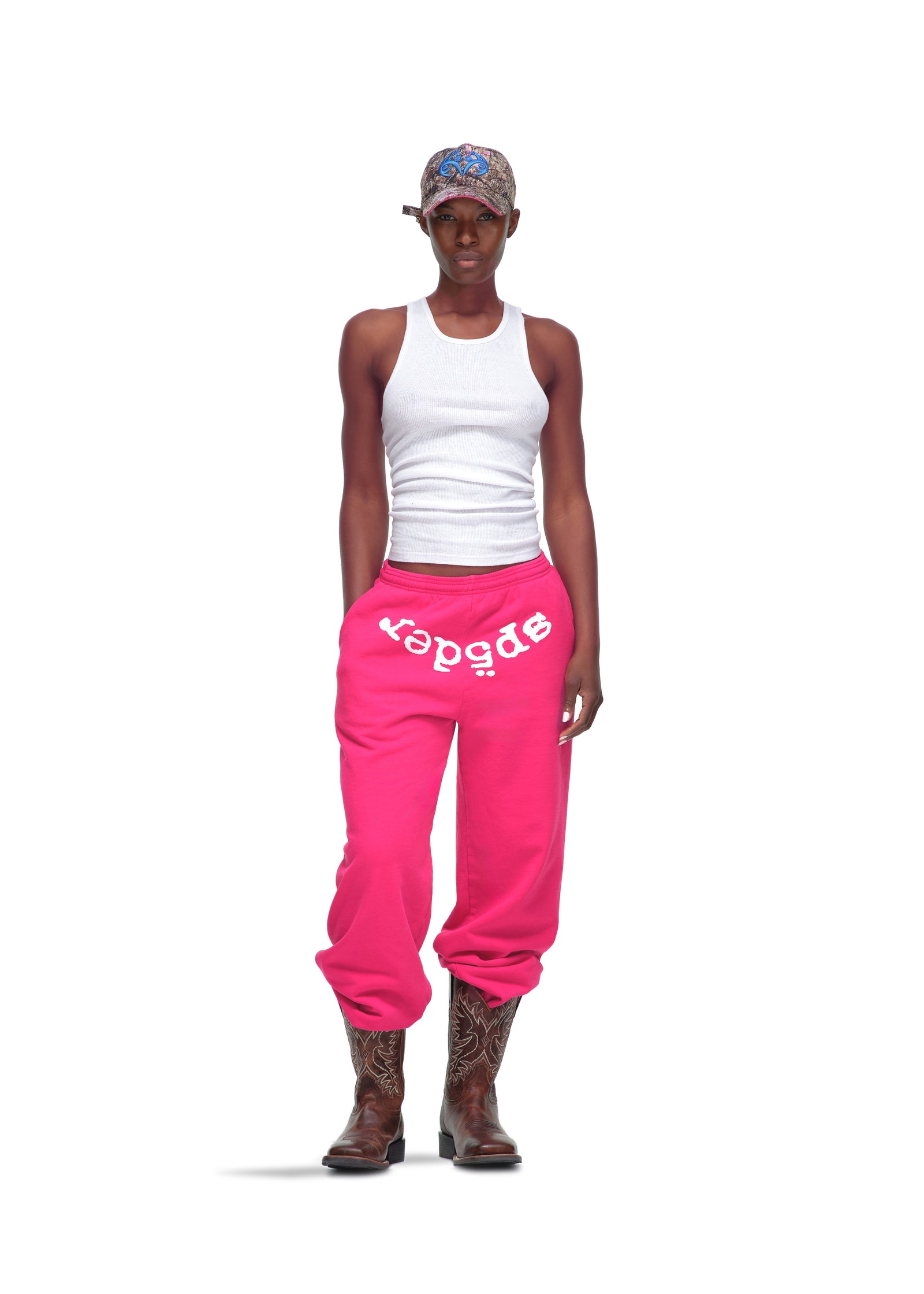 Female model in hot pink Legacy Sweatpants with white "SP5DER" puff print, white tank top, patterned cap, and cowboy boots.