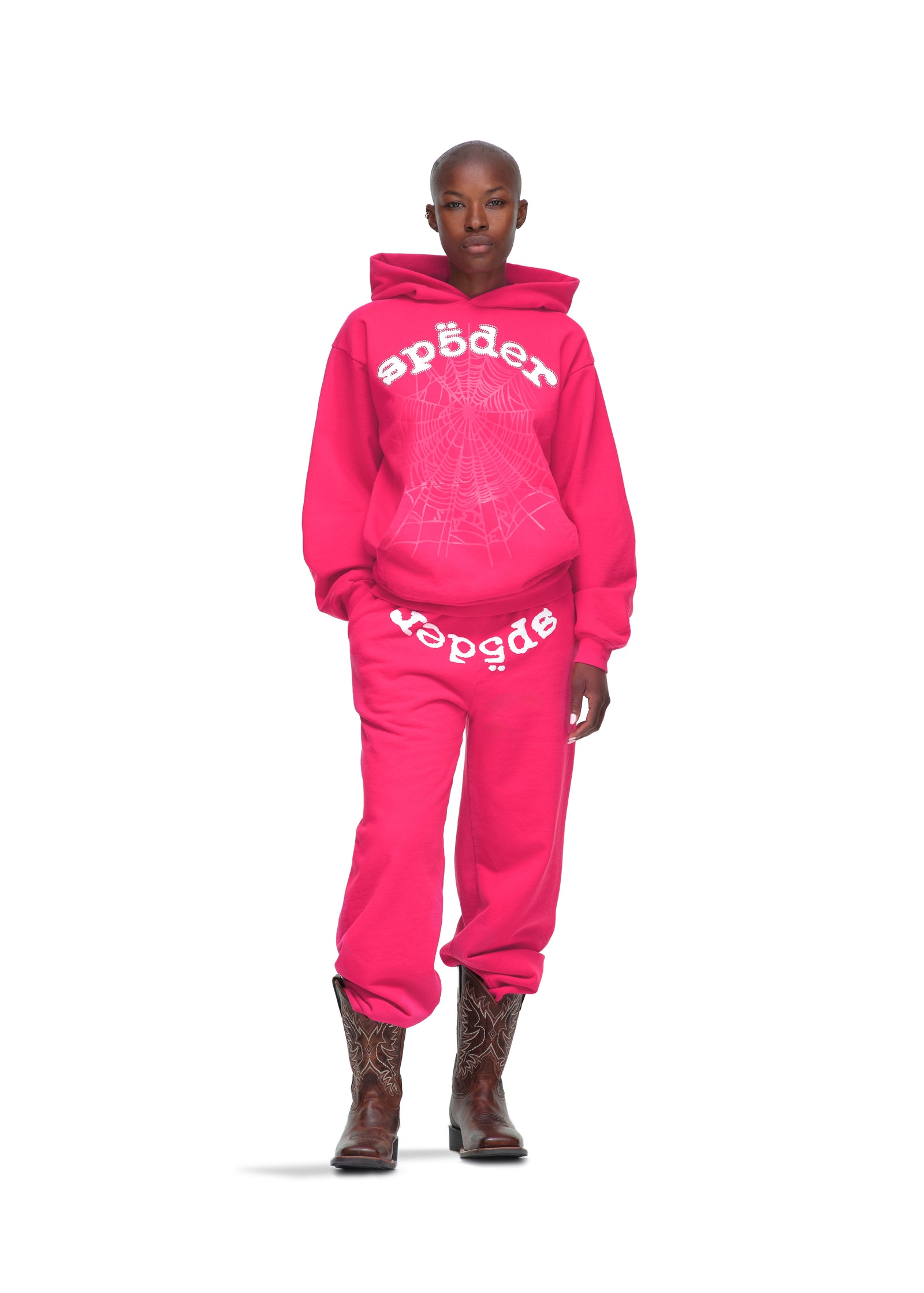 Female model in a pink Legacy Hoodie and sweatpants with white spider web and text print, wearing cowboy boots.
